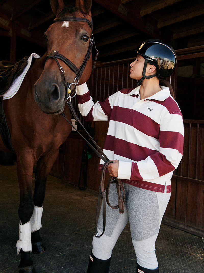 Model wearing a Club Polo in stripes, grooming a horse in a stable setting, showcasing comfort and style.