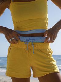 Woman wearing CloudKnit 3.5" Short CL in bright yellow with a blue top, on a beach backdrop.