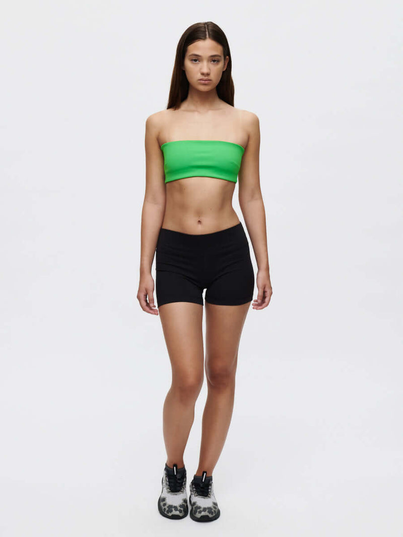 Model showcasing the Love Bra, a low-impact bandeau top in vibrant green, paired with black shorts and sporty shoes.