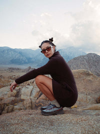 Model crouching on rocks wearing Boulder Dress in Dark Brown, with adjustable waist toggles and a cozy silhouette.
