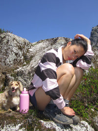 A woman crouches outdoors on rocky, grassy terrain under a blue sky, wearing a pink and black striped long-sleeve top, shorts, and hiking shoes. A small dog sits beside her next to a pink water bottle.