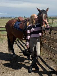 A woman in equestrian attire stands beside a brown horse in an outdoor paddock, kissing the horse's face while holding a black riding boot. The horse has a dark blue quilted saddle pad and a brown leather saddle.