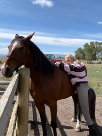 A woman in a pink and black striped long-sleeve shirt and grey breeches hugs a brown horse standing next to a wooden fence outdoors under a blue sky.