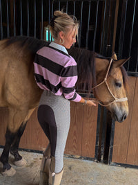 A woman in a pink and black striped long-sleeve shirt and gray riding breeches stands beside a brown horse in a stable, holding its reins.