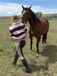 A woman in equestrian apparel stands beside a brown horse in an outdoor grassy field under a blue sky. The woman wears a pink and black striped cropped jacket, beige riding breeches with dark brown panels, and black riding boots.