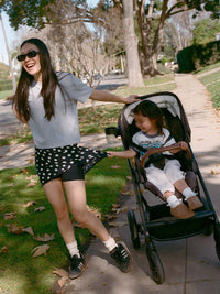 Woman smiling in a polka dot Club Skort, playing with a child in a stroller during a sunny day walk.