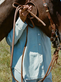 Person holding reins and wearing Desert Shirt in light blue, with oversized fit and brushed silver buttons.