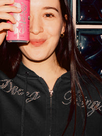 Model holding a drink and smiling while wearing The Diamanté Doing Things Hoodie in Black, featuring diamante script.
