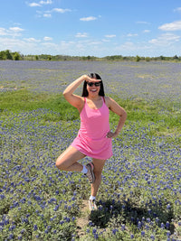 Model posing in The Exercise Dress in Pink with adjustable straps and built-in shorts liner, set in a field of bluebonnets.