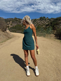 Model standing on a trail wearing The Exercise Dress in Teal with adjustable straps and built-in shorts liner.