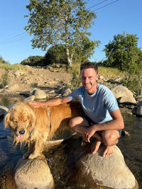Man kneeling by a stream wearing Everyday Classic Shortsleeve in Light Blue, enjoying outdoor time with his golden retriever.
