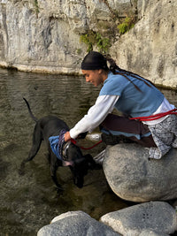 Woman kneeling on a rock, engaging with her dog by the water while wearing a blue Waffle Baby Tee in a natural outdoor setting.