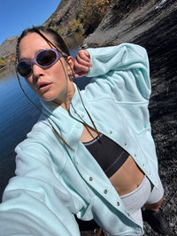 Model posing by the water in a light blue RecFleece Snap Jacket with dolman sleeves and snap-up front.