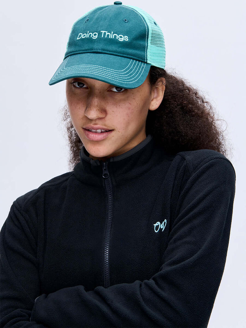 Model posing in Doing Things Trucker Hat in Teal, featuring breathable mesh back and embroidered logo.