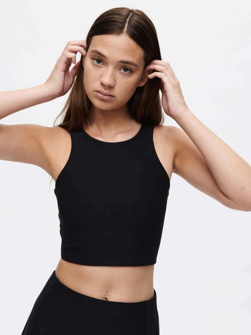 Model adjusting hair while wearing Venus Crop Top in Black, with the cropped silhouette and sleeveless design.