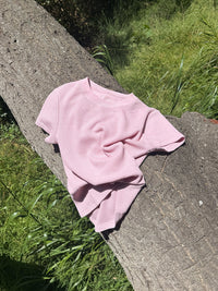 A light pink short-sleeve knit top with a textured waffle weave, draped on a large tree trunk outdoors with green grass in the background.