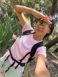 A woman wearing a light pink ribbed short-sleeve t-shirt, black hiking backpack straps, and sunglasses takes a selfie outdoors in a wooded area.