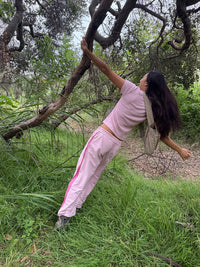 A woman posing outdoors in a grassy wooded area, wearing a light pink short-sleeve crop top and matching pink sweatpants with a pink stripe, holding onto a tree trunk.