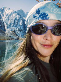 Smiling woman with long hair wearing sunglasses and a blue floral bandana, enjoying nature by the water.