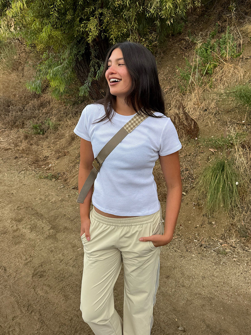 A woman standing outdoors on a dirt path wearing a white short-sleeve crop top, beige pants, and a beige plaid strap bag.