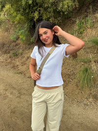 A young woman standing outdoors on a dirt path wearing a white short-sleeve crop top, beige track pants, and a beige crossbody bag with a plaid strap.