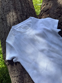 A white short-sleeve textured t-shirt with a fine mesh-like knit pattern, laid flat against a large tree trunk outdoors.