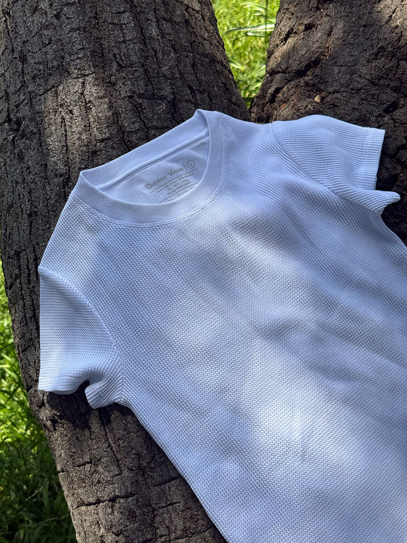 A white short-sleeve textured t-shirt with a fine mesh-like knit pattern, laid flat against a large tree trunk outdoors.