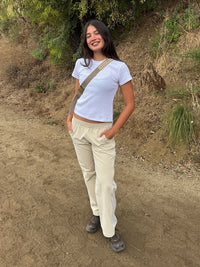 A woman stands on a dirt path outdoors wearing a white short-sleeve crop top, beige pants, and hiking shoes, with a beige crossbody bag across her body.